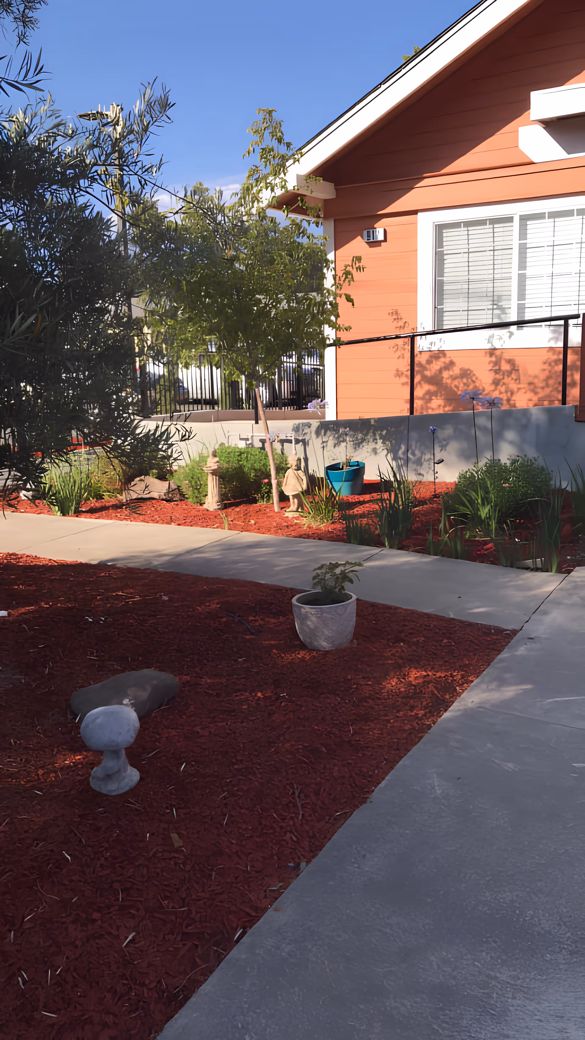 Outdoor garden area with a concrete pathway, red mulch, various plants, small statues, and a potted plant. A peach-colored building with white trim and a window with blinds is visible in the background under a clear blue sky.