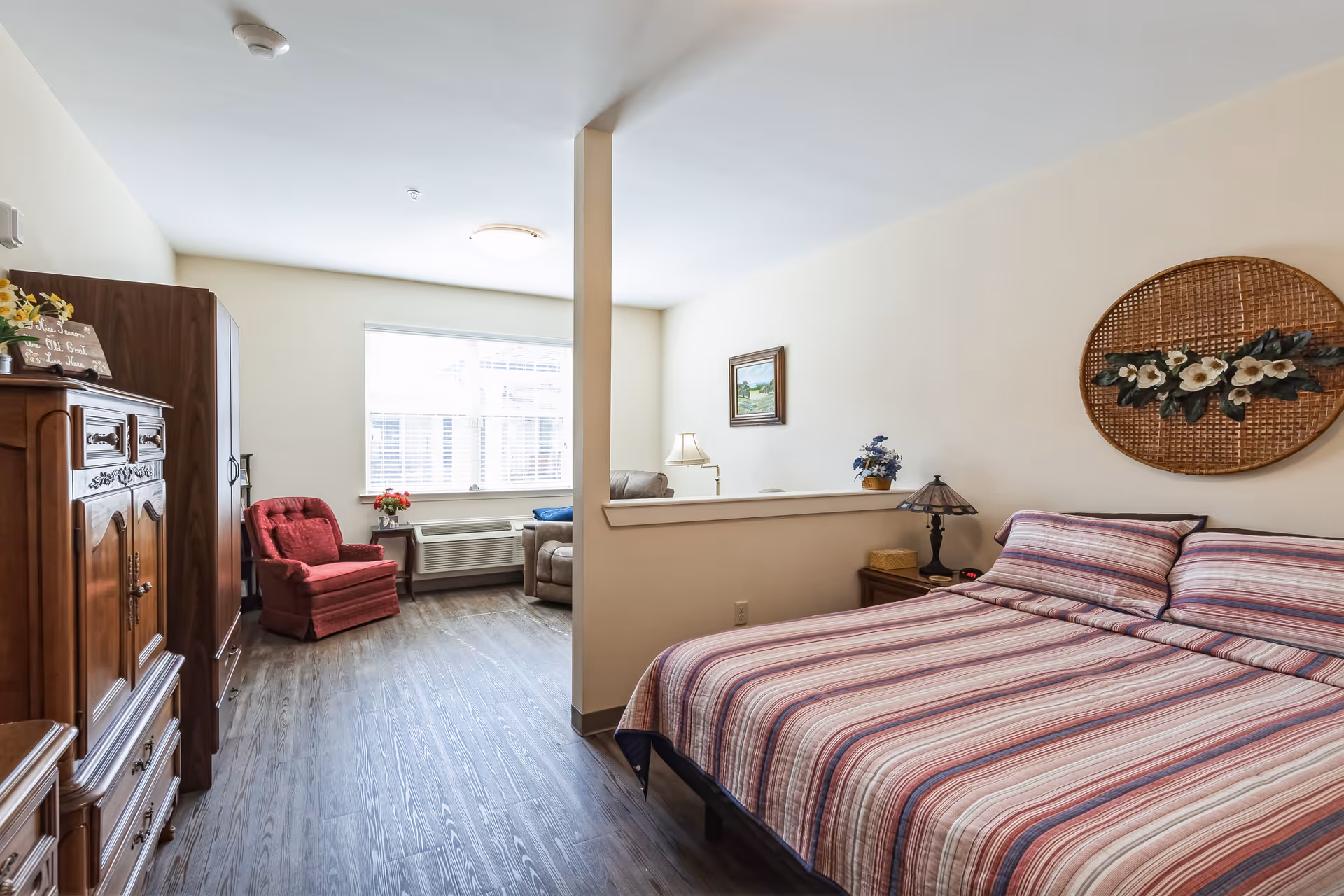 A bright, cozy bedroom with a large bed covered in a striped bedspread. The room features wooden furniture including a dresser and nightstand with a lamp. There is a partial wall separating the sleeping area from a sitting area with a red armchair, a beige recliner, and a window letting in natural light.