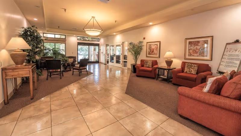 A spacious senior living facility common area with beige tiled floors and carpeted seating sections. The room features red upholstered armchairs and sofas with patterned cushions, wooden side tables with lamps, framed floral artwork on the walls, and large windows and glass doors letting in natural light. There are also green plants adding a touch of nature to the space.