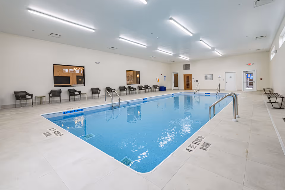 Indoor swimming pool with clear blue water surrounded by beige tiled flooring. Several black chairs and small tables are lined up against the white walls. The room is well-lit with multiple ceiling lights, and there are windows and doors visible on the walls.