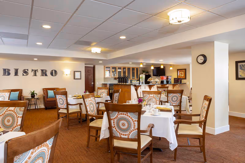 A cozy dining area with several tables covered in white tablecloths, each set with cups, glasses, and napkins. Wooden chairs with patterned cushions surround the tables. The wall in the background has the word 'BISTRO' displayed, and there is a counter area with coffee dispensers and a television mounted above. The room has warm lighting and a carpeted floor.