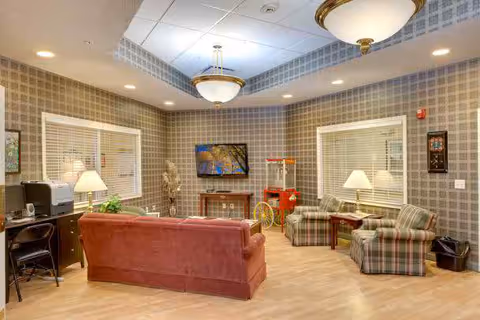 Well-lit communal lounge with a sofa and plaid armchairs facing a wall-mounted TV and side tables.