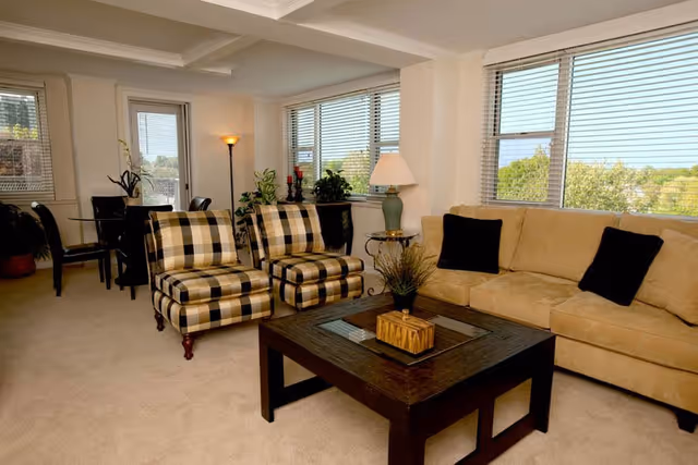 A bright living room with large windows showing a view of trees outside. The room features a beige sofa with two black pillows, two plaid armchairs, a dark wooden coffee table with decorative items, a side table with a lamp, and a dining area with a glass table and black chairs in the background.