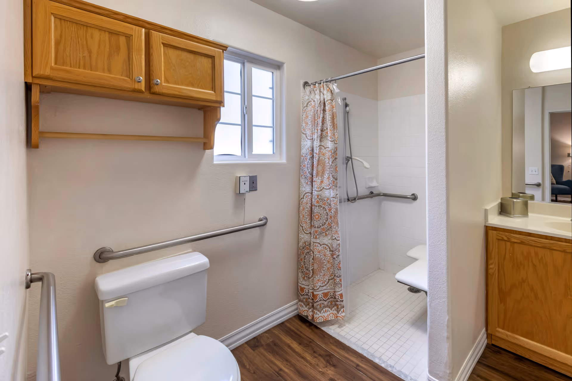 A bathroom with a white toilet, wooden cabinet mounted on the wall, a window, and a walk-in shower with a patterned shower curtain and grab bars. There is a wooden vanity with a sink and a mirror on the right side.