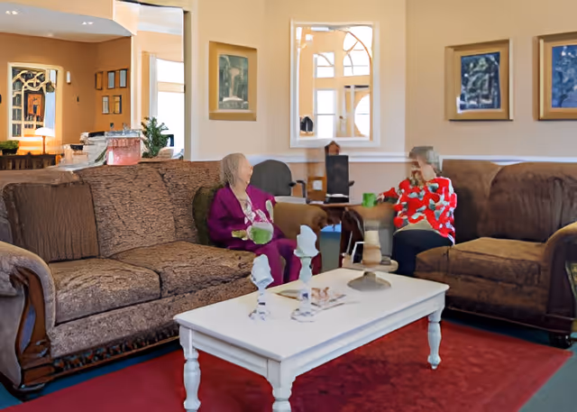 A cozy living room with two elderly women sitting on separate brown sofas facing each other. A white coffee table with decorative items is in the center on a red rug. The walls have framed artwork and a mirror, and there is a warm, inviting atmosphere.