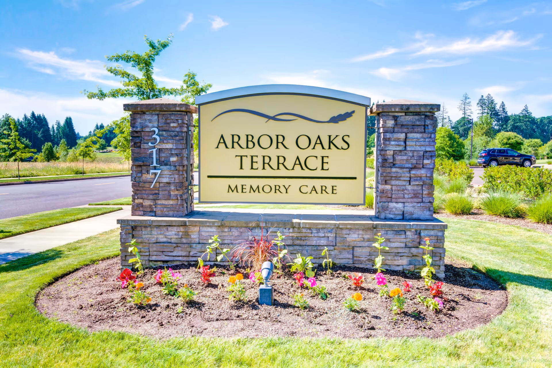Stone sign for Arbor Oaks Terrace Memory Care Residence with the address number 317 on a pillar, surrounded by a flower bed with colorful flowers and green grass, under a blue sky with some clouds.