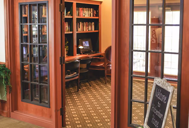 Interior view of a cozy room with wooden framed glass doors partially open, revealing a small workspace with a computer, bookshelves filled with books, and chairs. A decorative carpet covers the floor, and a chalkboard sign near the door reads 'Welcome to Habersham House.'