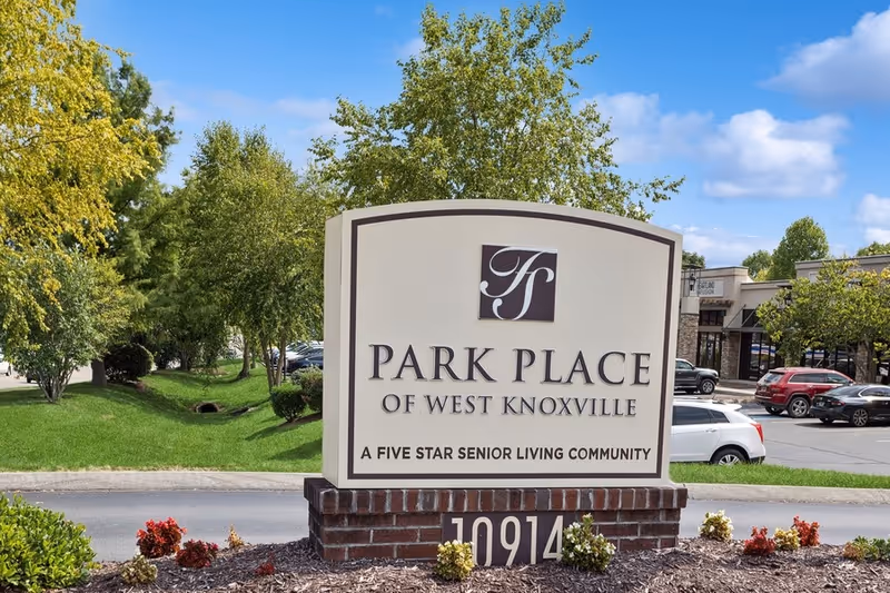 Monument entrance sign reading "Park Place of West Knoxville — A Five Star Senior Living Community" set on landscaped grounds with parked cars and buildings behind it.