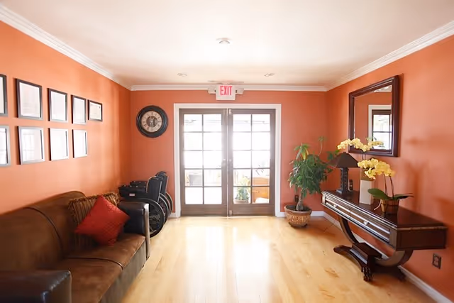 A bright living room with orange walls, a brown leather sofa with red and brown cushions on the left, a set of framed pictures and a clock on the wall above the sofa, and a wooden console table with a lamp, yellow orchids, and a large mirror on the right. There is a potted plant next to the console table and double glass doors with an exit sign above them at the far end of the room.