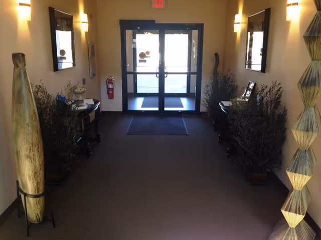 Interior view of a hallway leading to a glass double door exit. The hallway is carpeted and decorated with plants, two small tables with decorative items, wall-mounted lights, and mirrors on both sides.