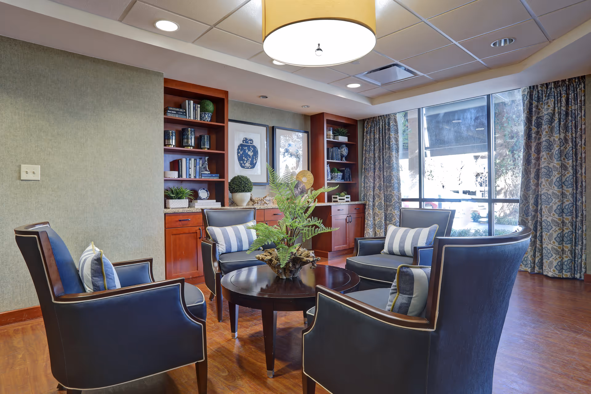 A cozy sitting area with four dark blue armchairs arranged around a round wooden coffee table with a plant centerpiece. Behind the chairs, there are built-in wooden shelves with decorative items and framed artwork on the wall. Large windows with patterned curtains let in natural light.