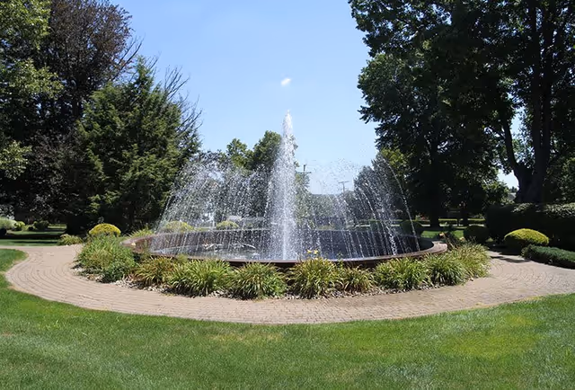 A circular water fountain surrounded by green grass, plants, and trees under a clear blue sky in an outdoor garden area.