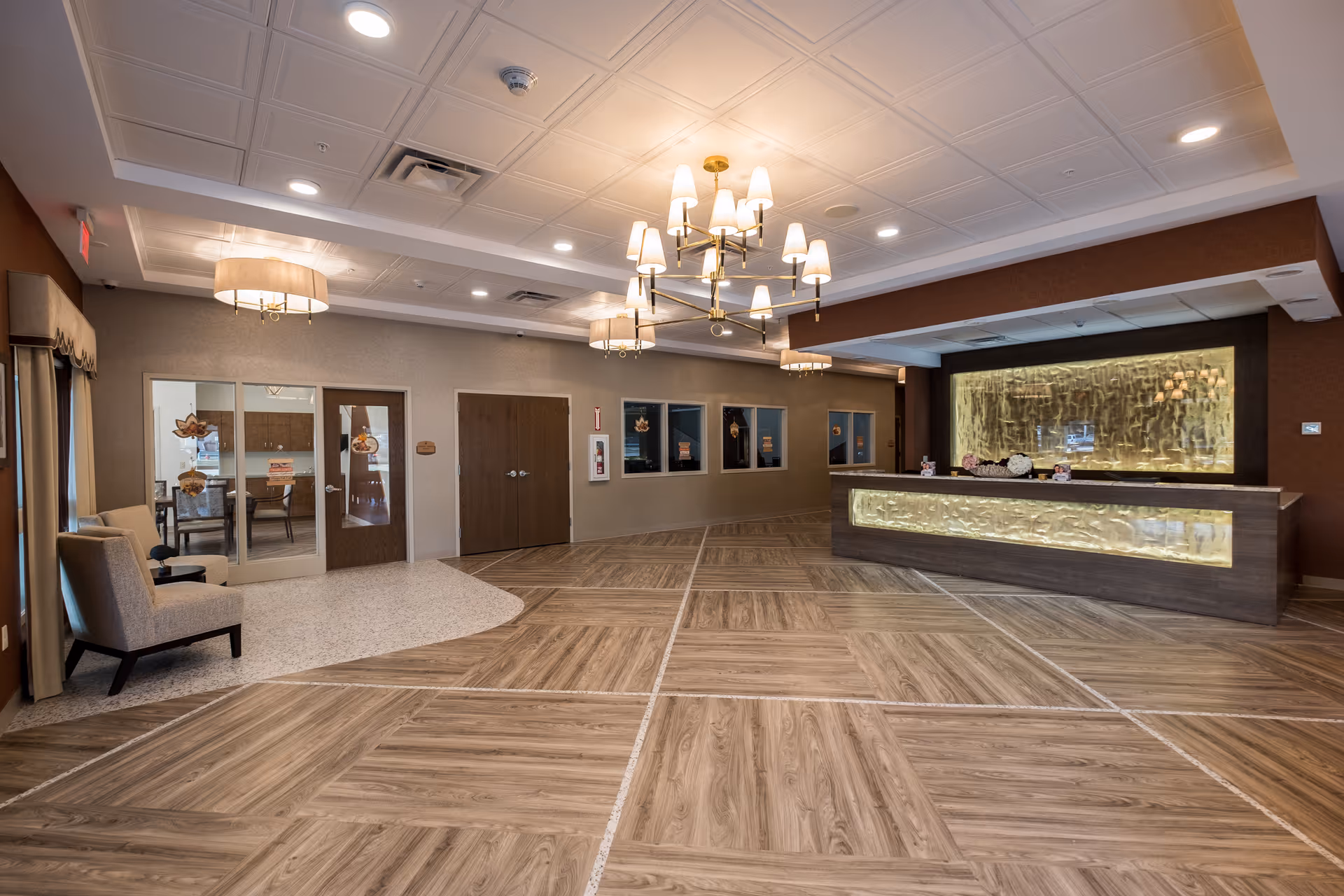 Spacious and well-lit reception area with a modern chandelier, a large wooden reception desk with illuminated panels, wood-patterned flooring, beige walls, and comfortable seating near glass doors leading to another room.