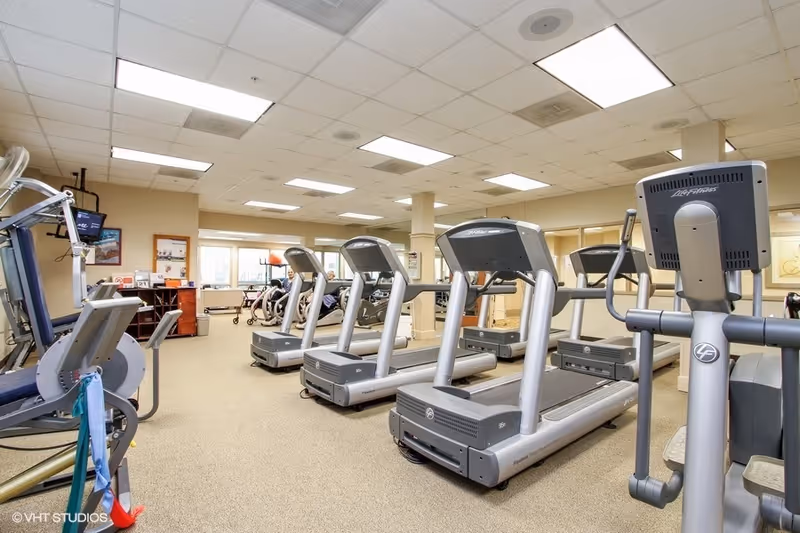 A fitness room with several treadmills and exercise machines arranged in rows. The room has a beige carpet, white ceiling tiles with fluorescent lights, and large windows letting in natural light. There is a small desk and shelving unit in the back corner with some equipment and a walker visible.