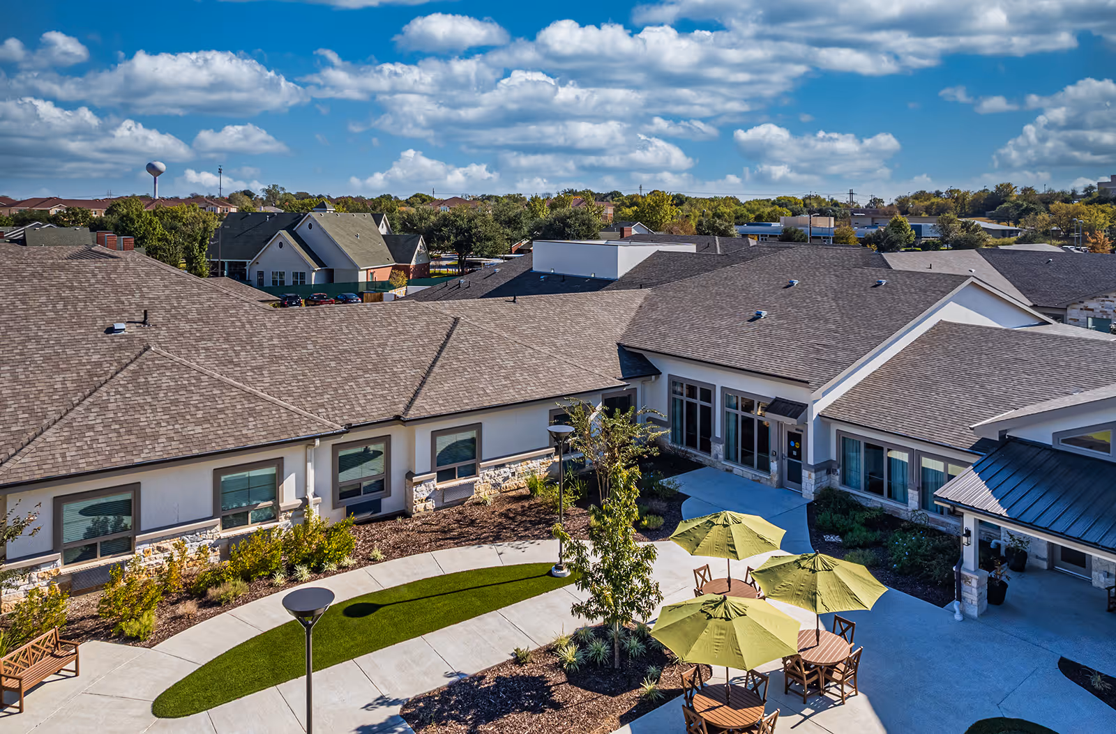 Aerial view of a senior living facility courtyard with tables, green umbrellas, landscaping and surrounding single-story buildings under a blue sky.