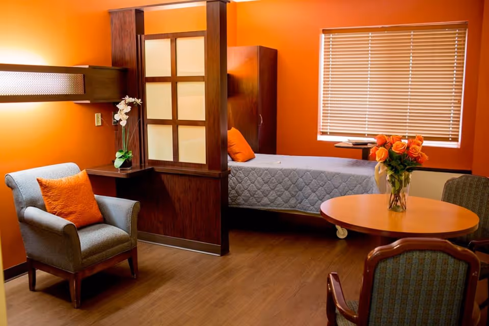 A cozy room with orange walls featuring a single bed with a gray quilt and an orange pillow, a gray armchair with an orange cushion, a round wooden table with a vase of orange roses, and two chairs. There is a window with closed blinds and a wooden partition with frosted glass panels separating the bed area from the seating area.