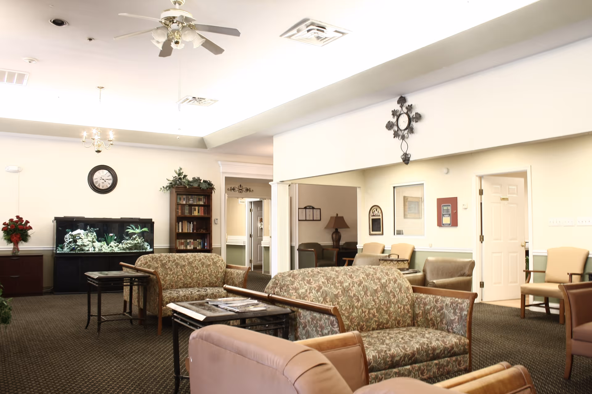 Well-lit senior living lounge with floral-patterned sofas and armchairs, a large fish tank, bookshelf, and ceiling fans.
