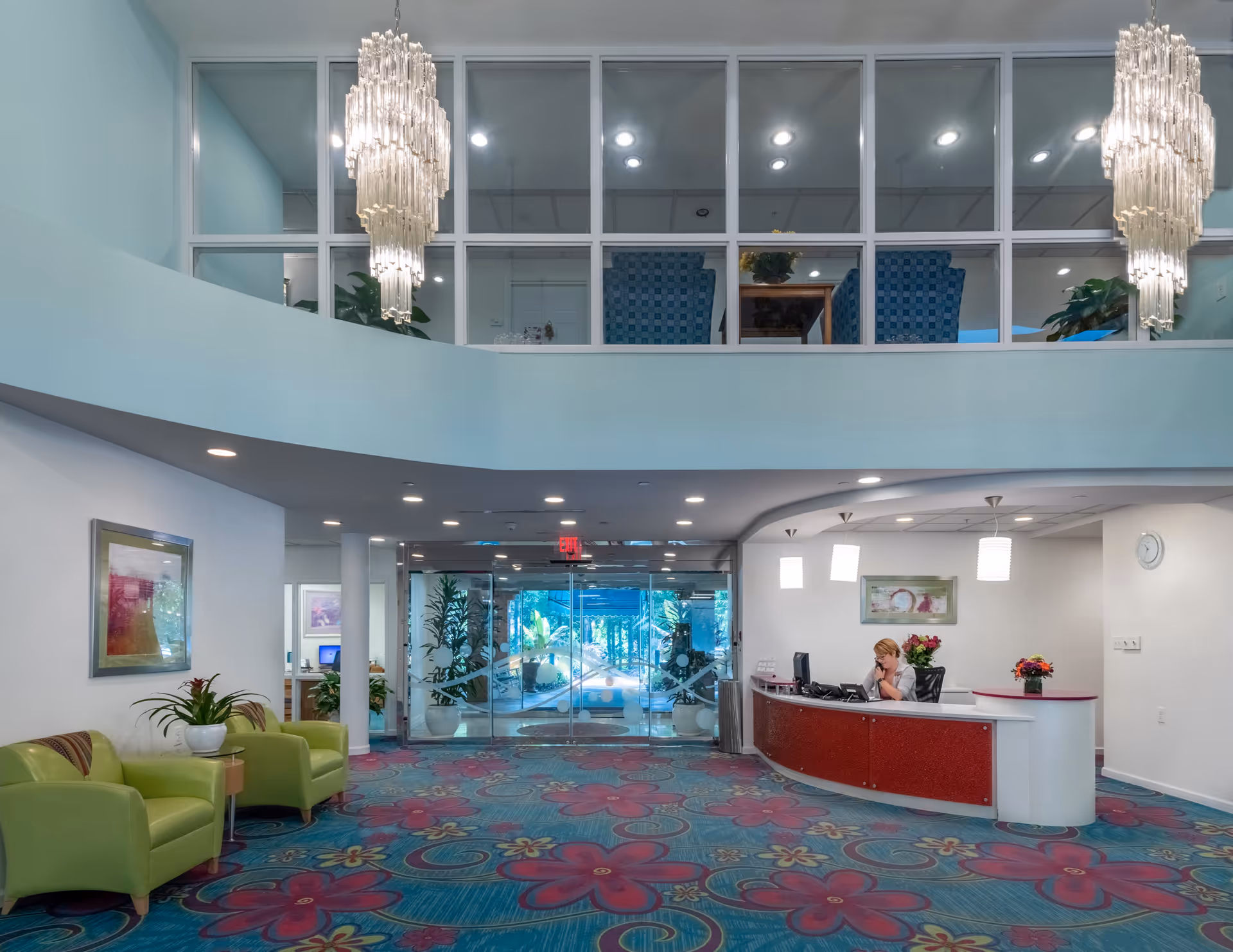 Reception area of Delmar Gardens of Smyrna featuring a curved red and white reception desk with a receptionist seated behind it. The floor is covered with a colorful carpet with large pink and red floral patterns. Two green armchairs with a small table and potted plant are on the left side. Above, there are large windows showing a second-floor seating area with blue chairs and two large chandeliers hanging from the ceiling. Glass doors lead to an outdoor area with plants visible through the entrance.