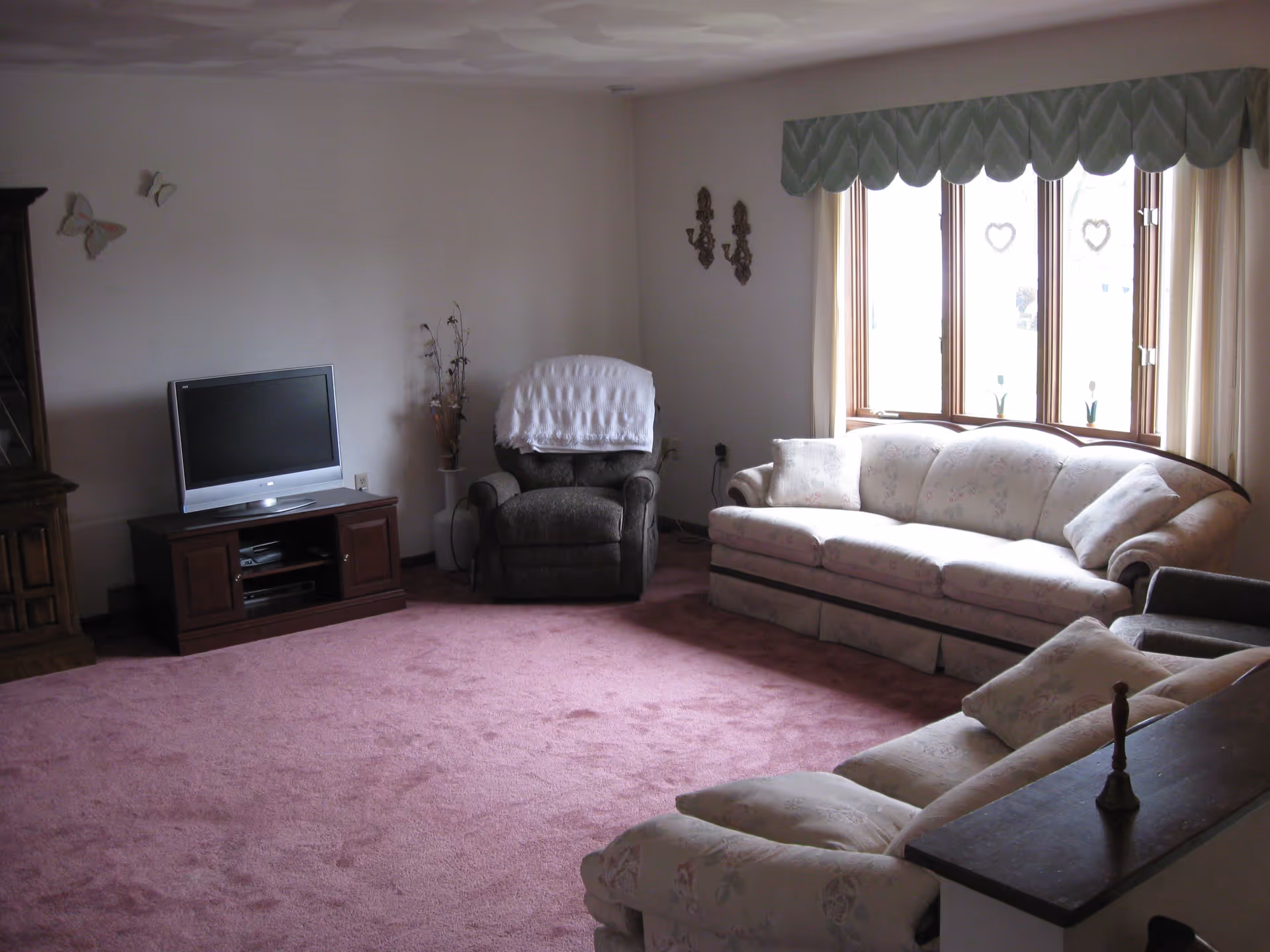 A bright living room with floral sofas, an armchair, a TV on a wooden stand, and a large bay window over pink carpeting.