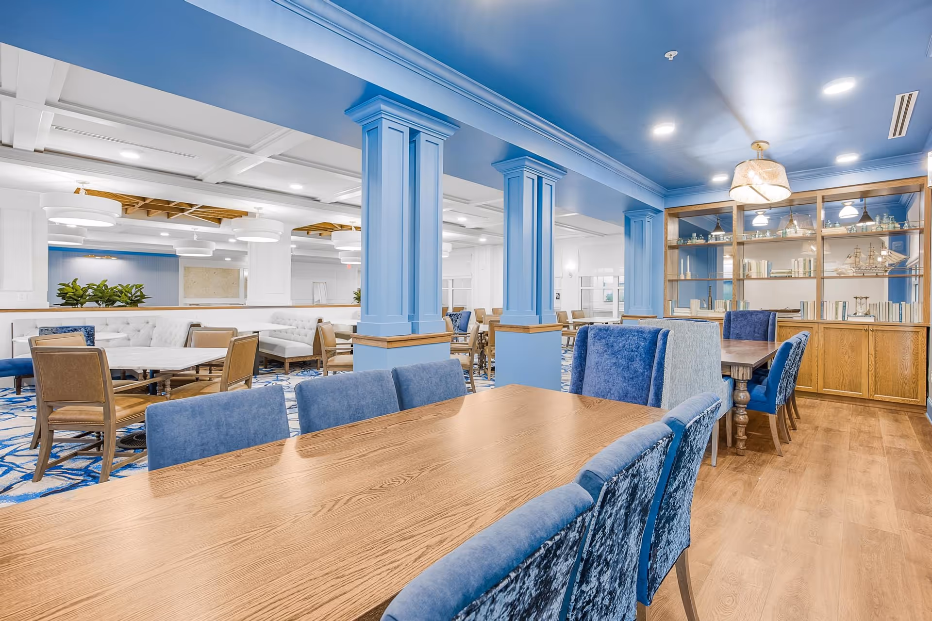 A spacious and elegant dining area in a senior living facility featuring a long wooden table with blue upholstered chairs in the foreground. The room has blue pillars, a wooden floor, and a built-in wooden shelving unit with decorative items and books. Additional seating areas with tables and chairs are visible in the background, along with white tufted benches and modern ceiling lights.