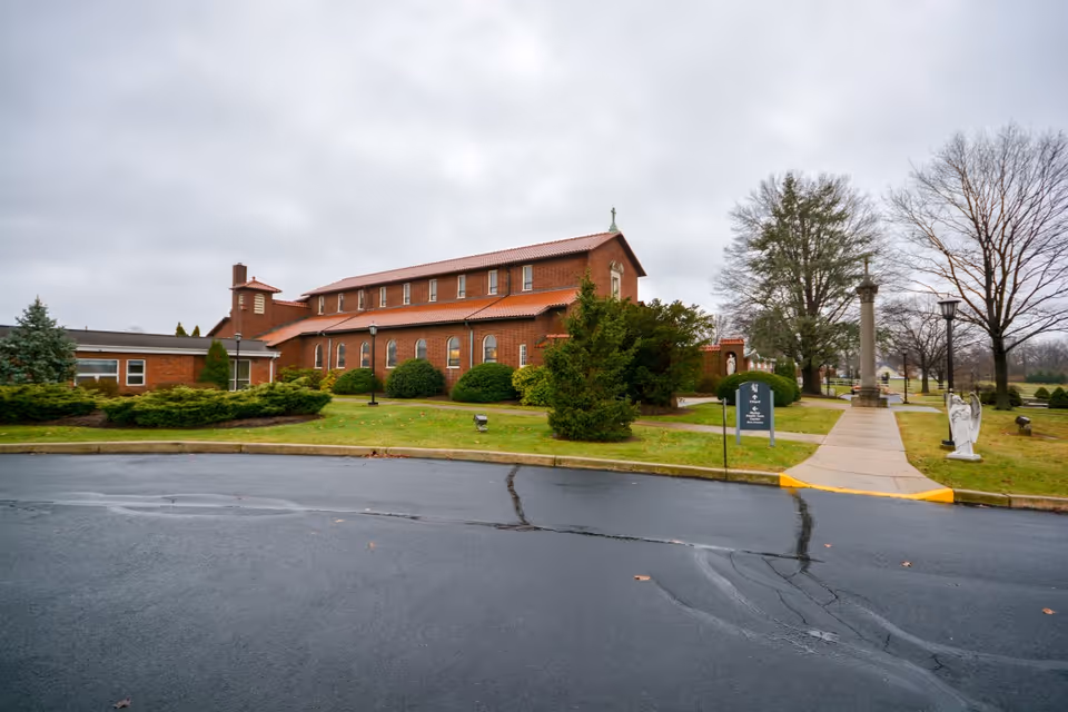 Exterior view of a brick building with a red tiled roof, surrounded by green bushes and trees on a cloudy day. A paved walkway leads to the building entrance, with a statue of an angel and a tall monument visible on the right side of the walkway.