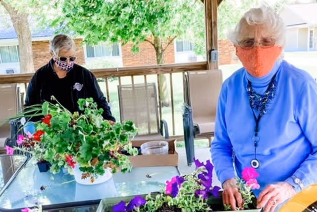 Two elderly women wearing face masks are tending to potted flowering plants on a glass table in a covered outdoor patio area with chairs and trees visible in the background.