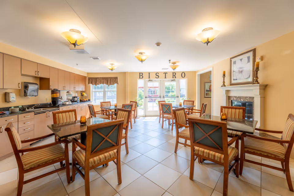 A bright and inviting bistro dining area with several wooden tables and chairs featuring striped cushions. The room has a tiled floor, warm yellow walls, and ceiling lights. There is a fireplace with decorative candles and a framed flower picture above it on the right wall. On the left side, there is a counter with cabinets, a coffee machine, and other beverage dispensers. Large windows and glass doors at the far end let in natural light and have the word 'BISTRO' displayed above them.