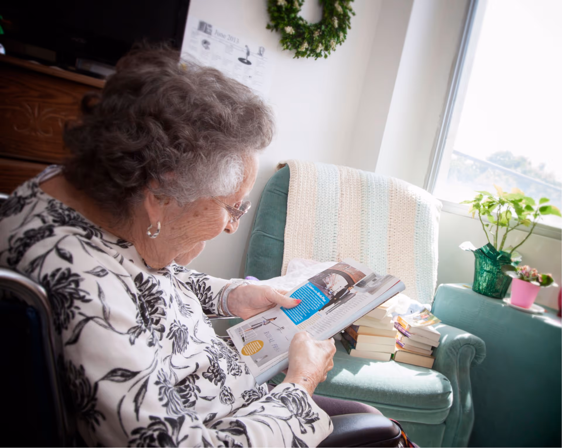An elderly woman with curly gray hair and glasses is sitting in a wheelchair, reading a book in a cozy room. Behind her is a green armchair with a light-colored knitted blanket draped over it, a stack of books on the seat, and two potted plants on a nearby table by a window letting in natural light.