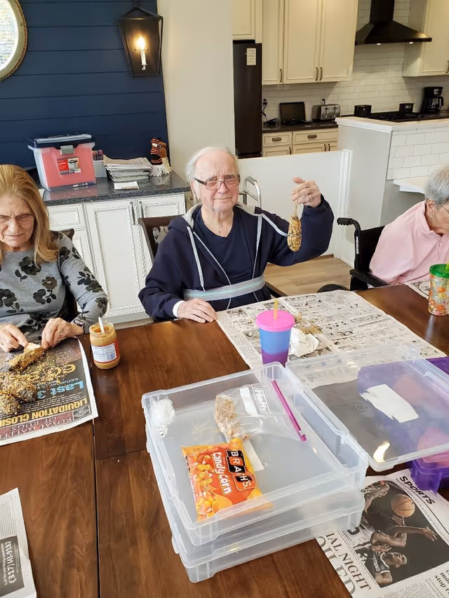 Three elderly individuals sitting around a wooden table in a kitchen or dining area. The man in the center is holding up a homemade bird feeder made of seeds. The woman on the left is working with peanut butter and seeds, and the woman on the right is seated with a green cup. The background shows white cabinets, a refrigerator, and a stove with a range hood.