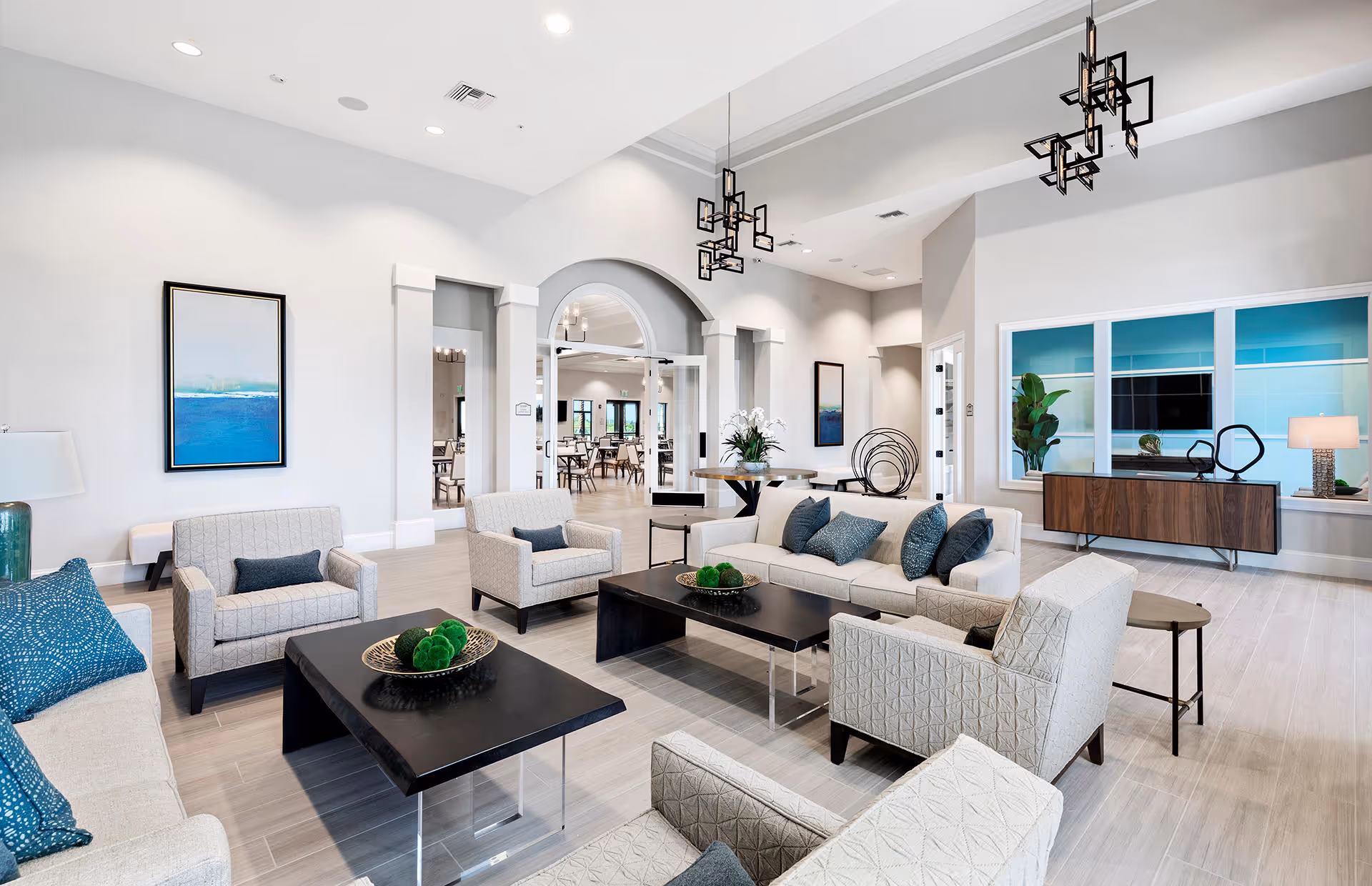 Bright and spacious living room area in a retirement community featuring multiple beige armchairs and sofas with blue and gray cushions arranged around two black coffee tables. The room has light-colored flooring, modern geometric pendant lights, a large window with a view into another room, and decorative elements including framed artwork, plants, and a sideboard.