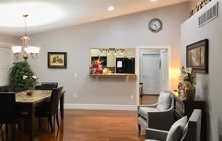 Interior view of a senior living facility dining and living area with a dining table and chairs on the left, two armchairs on the right, a pass-through window to the kitchen, wall clock above the doorway, and decorative plants and framed pictures on the walls.