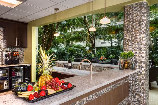 Indoor kitchen area with a large granite countertop island featuring a sink and a tray of fresh fruits including pineapple, grapes, and strawberries. The kitchen has dark wood cabinets, a wine cooler, and a coffee machine. Large windows behind the island show lush green plants and trees outside. Pendant lights hang above the island.