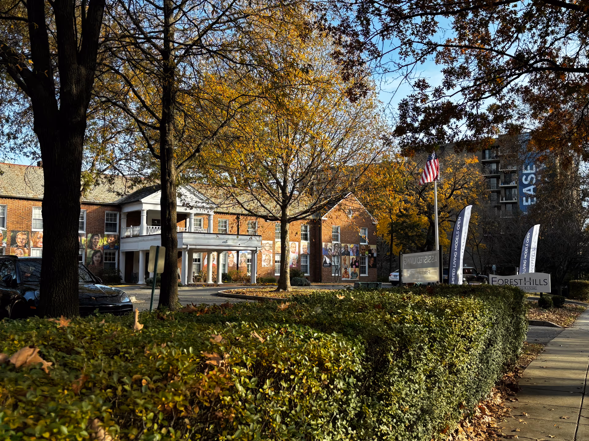Exterior view of a senior living facility named Forest Hills, showing a brick building with white columns at the entrance, surrounded by trees with autumn foliage and green hedges. There are flags and signs near the sidewalk, and a few parked cars are visible.