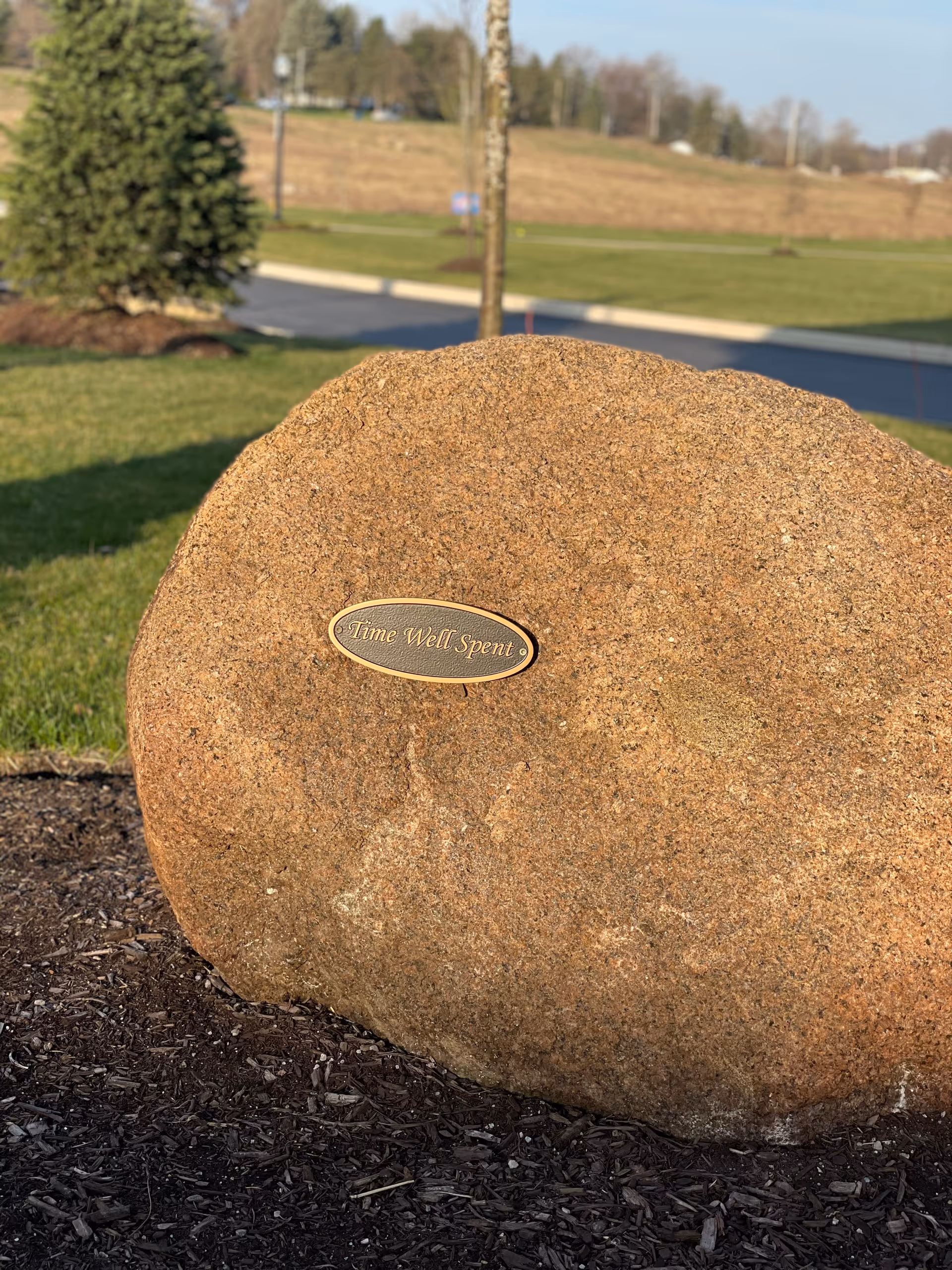 A large brown rock outdoors with a small oval plaque attached to it that reads 'Time Well Spent'. The rock is surrounded by mulch and grass, with a tree and a paved road visible in the background under a clear sky.