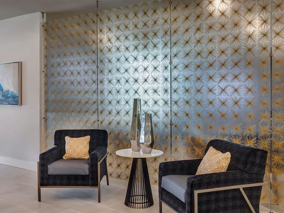 Two black armchairs with gray cushions and yellow patterned pillows flank a small round table with two tall metallic vases, set against a decorative glass wall with a gold starburst pattern in a modern interior space.