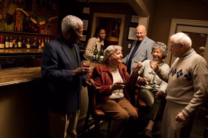 A group of five elderly people socializing and enjoying drinks together in a cozy bar area, with a bartender standing behind the counter smiling.