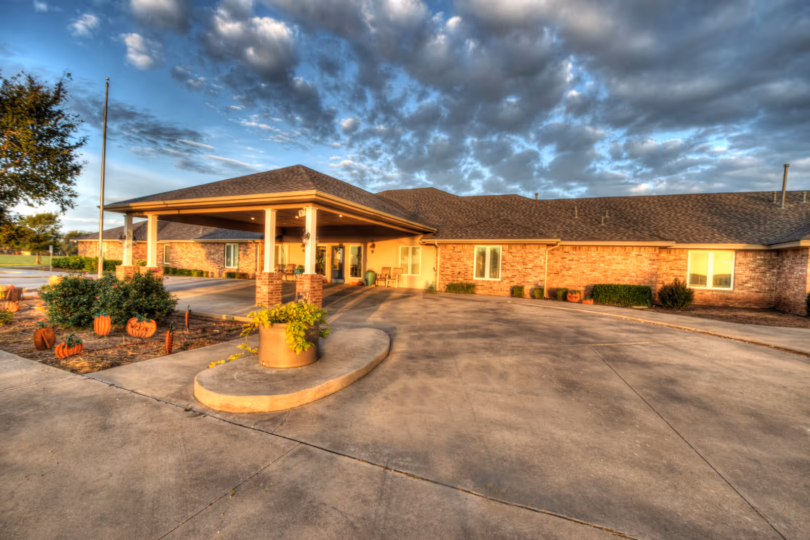 Exterior view of a single-story brick building with a covered entrance and a driveway. There are some plants and Halloween-themed pumpkin decorations near the entrance. The sky is partly cloudy with a warm light illuminating the scene.