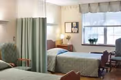 Two hospital-style beds with a privacy curtain, nightstand, desk, and window in a resident bedroom.