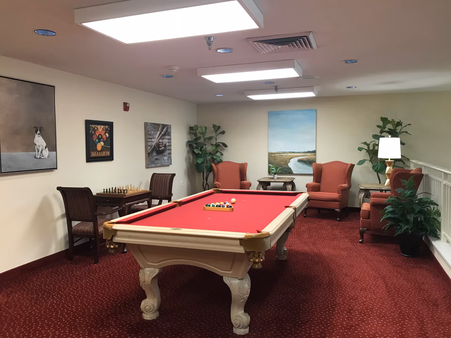 A senior living facility game room with a red felt pool table in the center, surrounded by four red upholstered armchairs and side tables with lamps. The room has red carpet, beige walls adorned with paintings, and a small table with two chairs and a chessboard. There are also green plants adding a touch of nature to the space.