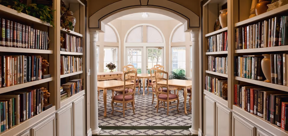 View through an archway into a bright room with wooden tables and chairs with purple cushions. The room has large windows with arched transoms letting in natural light. On either side of the archway are built-in bookshelves filled with books and decorative vases.