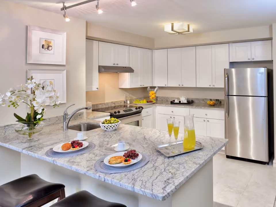 Bright modern kitchen with a marble island, stainless steel refrigerator, white cabinets, and place settings with drinks.