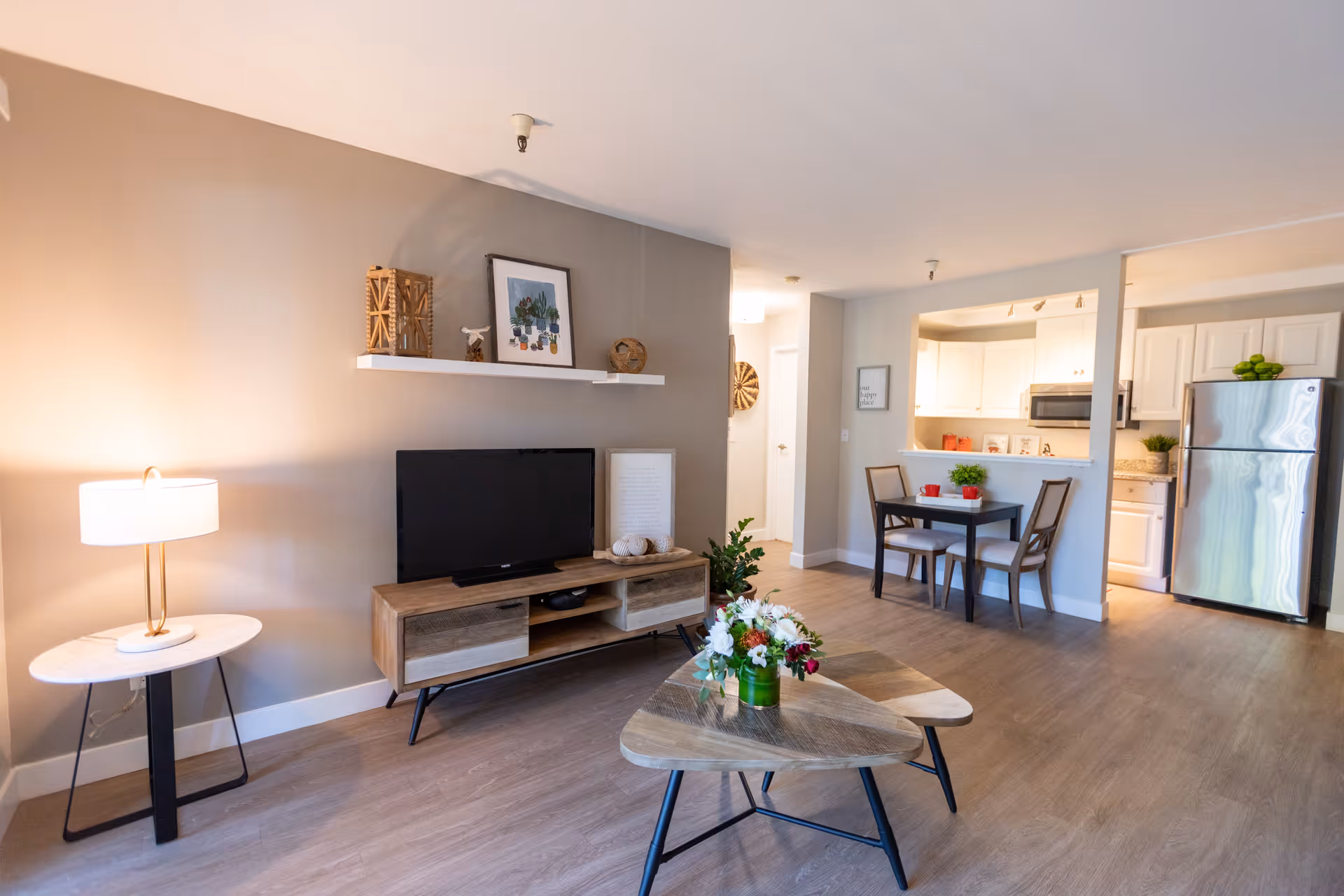 A bright and modern senior living apartment interior featuring a living room with a wooden TV stand, flat-screen TV, side table with a lamp, and a coffee table with a flower arrangement. In the background, there is a small dining area with a table and two chairs, and a kitchen with white cabinets, a microwave, and a stainless steel refrigerator.