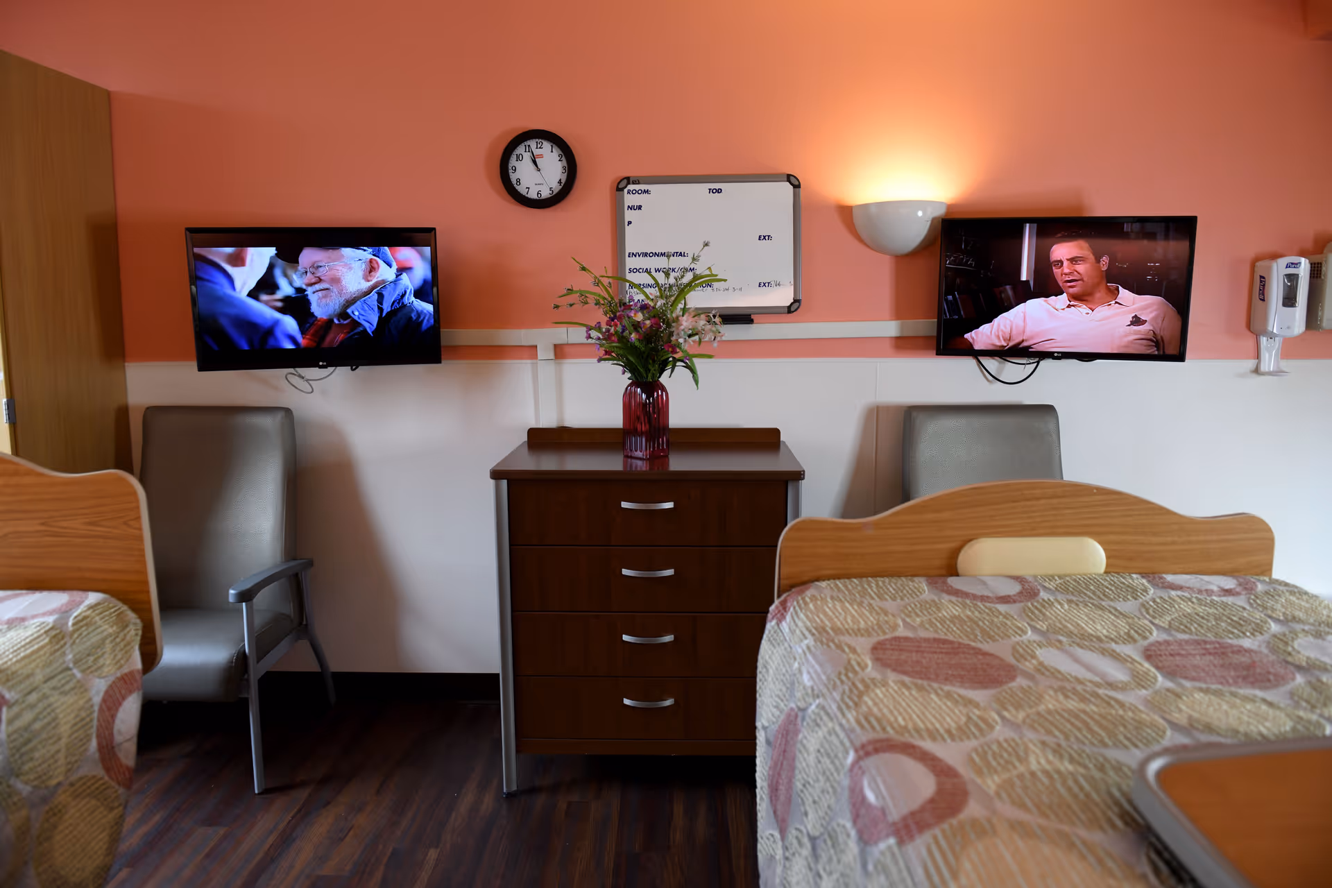 A shared bedroom in a senior living facility with two beds covered in patterned bedspreads, two gray chairs, a wooden dresser with a vase of flowers, a wall clock, a whiteboard, and two wall-mounted televisions showing different programs.