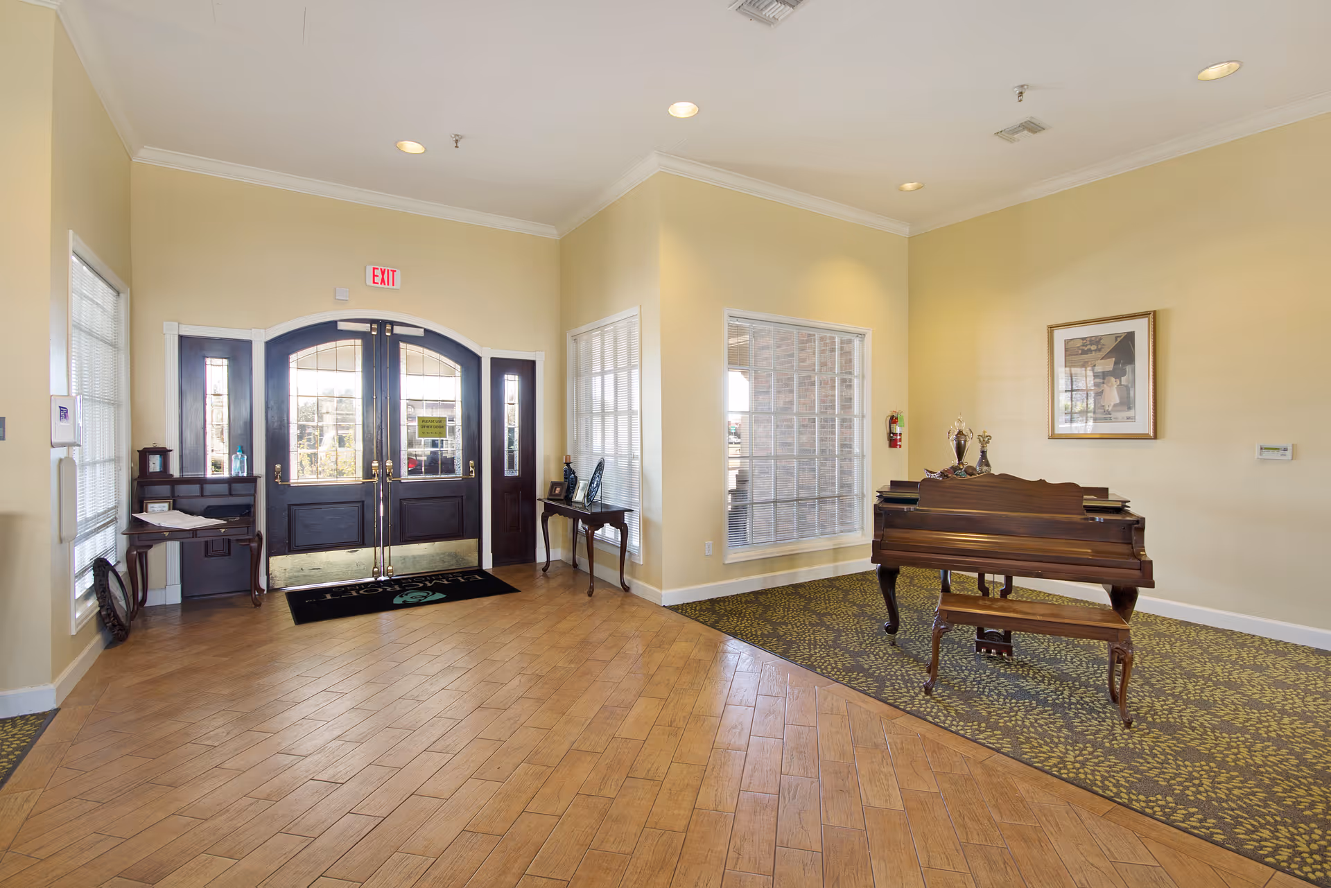 Interior view of a senior living facility entrance area with double dark wooden doors featuring glass panels. The floor is a combination of wood and patterned carpet. To the right, there is a wooden piano with a matching bench, decorative items on top, and a framed picture on the wall above. On the left side, there are two small tables with decorative items and windows with blinds.