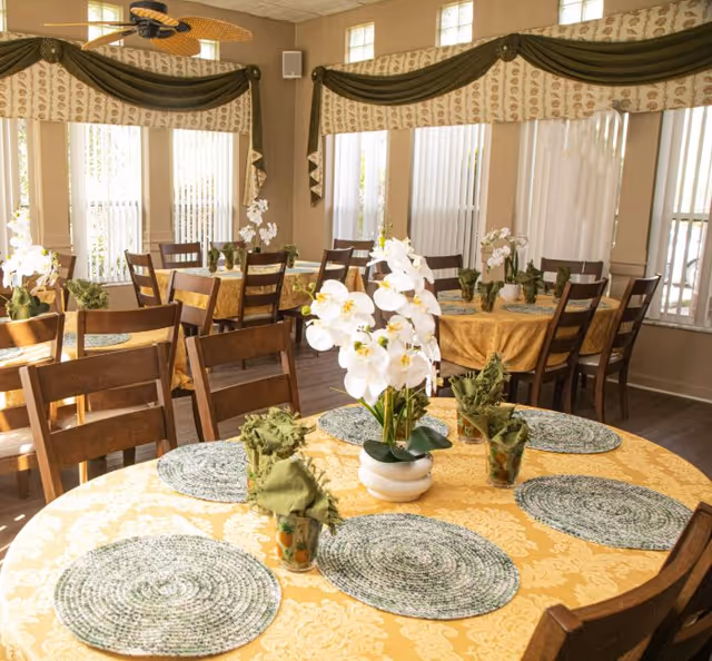 A bright dining room with multiple round tables covered in yellow patterned tablecloths. Each table has green woven placemats, green cloth napkins in glass holders, and white orchid centerpieces. Wooden chairs surround the tables, and large windows with vertical blinds and decorative valances allow natural light to fill the room.