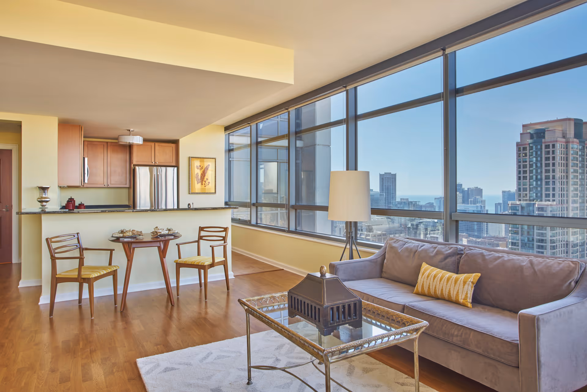 A modern living area with large floor-to-ceiling windows offering a cityscape view. The room features a gray sofa with a yellow patterned pillow, a glass coffee table with a decorative item, a floor lamp, and a small dining table with two wooden chairs. The kitchen area has wooden cabinets, a stainless steel refrigerator, and a countertop with some decorative items.