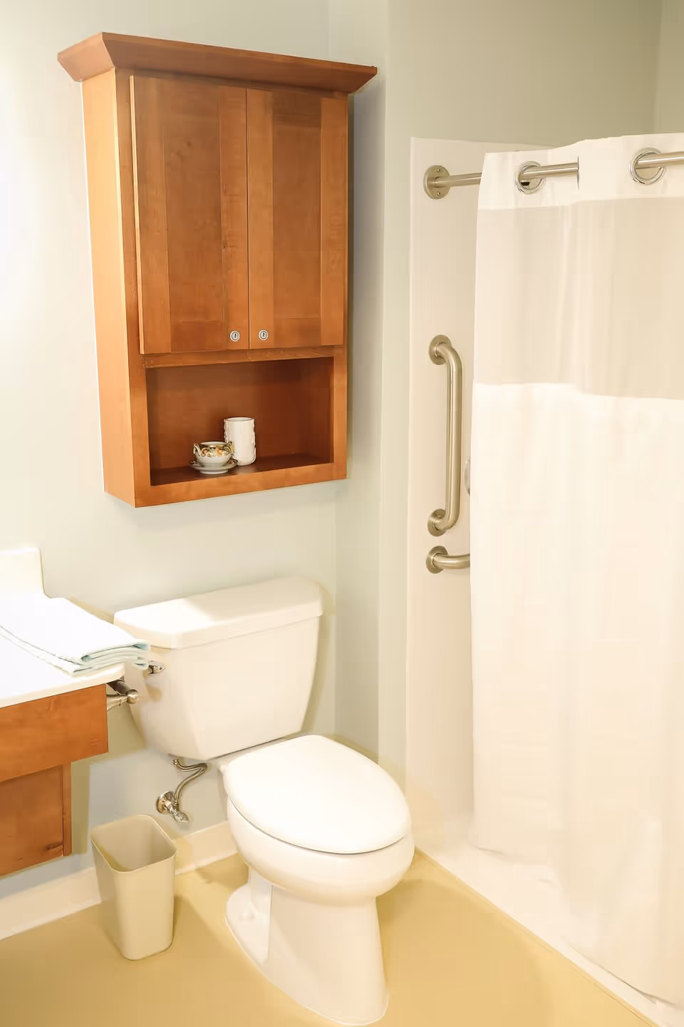A clean bathroom featuring a white toilet, a wooden cabinet mounted on the wall above the toilet with two closed doors and an open shelf holding a cup and a small decorative item. To the right is a shower area with a white curtain and metal grab bars. A small trash bin is placed on the floor next to the toilet.