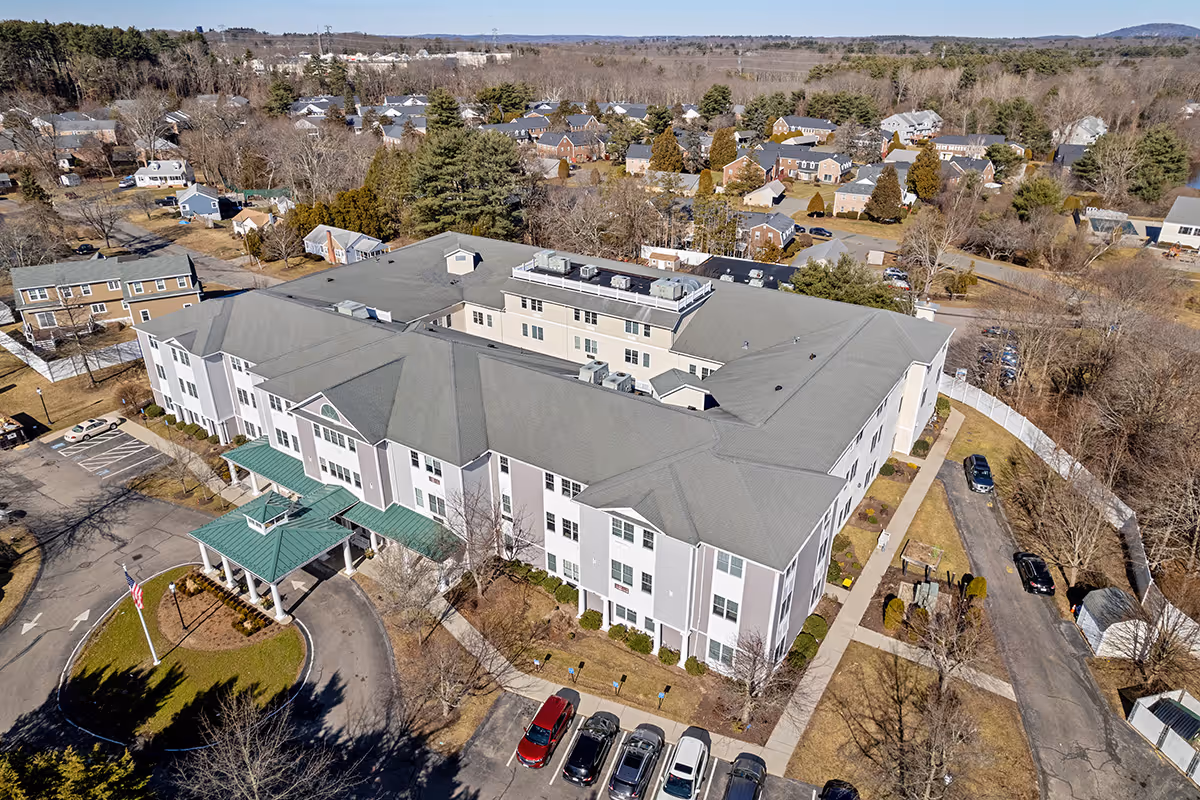 Aerial view of a large senior living facility building with a green entrance canopy and surrounding parking spaces. The building is three stories tall with a gray roof and white exterior walls. The facility is located in a suburban neighborhood with many houses and trees visible in the background.