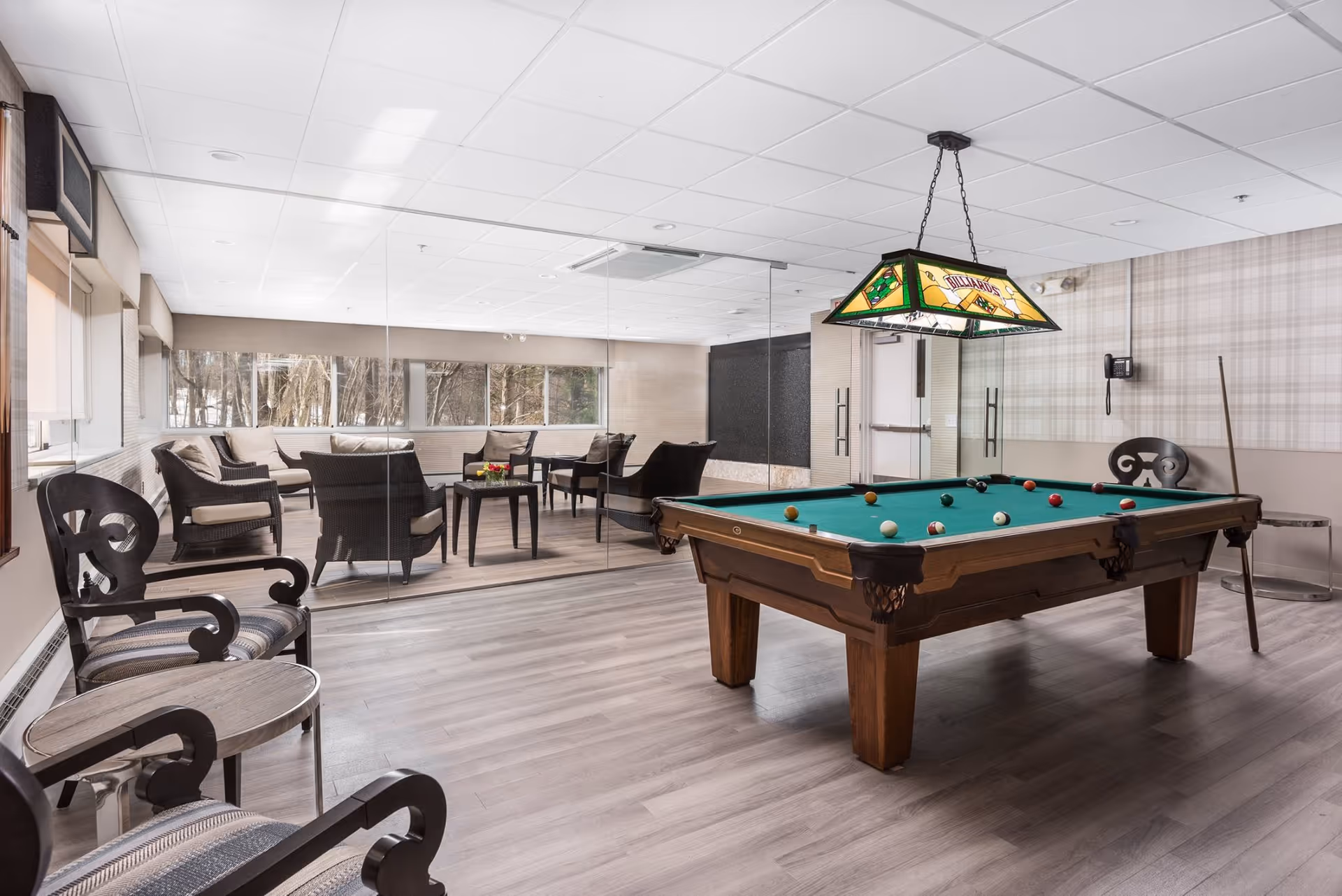 A bright indoor common room featuring a pool table with a stained-glass hanging light and a seating area of chairs and tables by large windows.