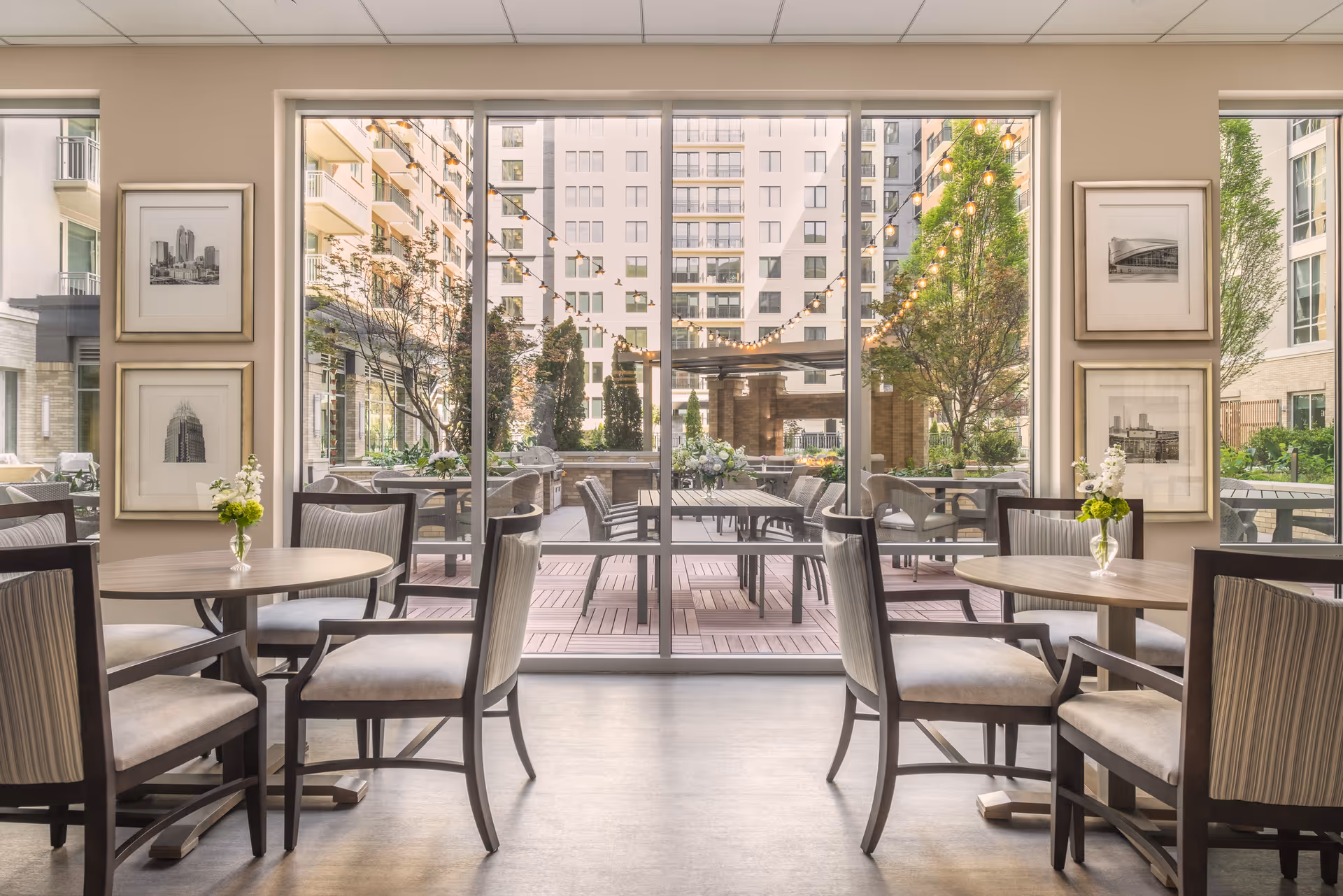 Interior dining area with round tables and cushioned chairs, each table decorated with a small vase of flowers. Large floor-to-ceiling windows provide a view of an outdoor patio with additional seating, string lights, trees, and surrounding buildings.